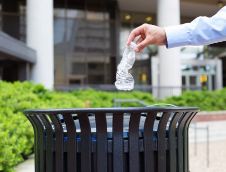 A person's hand, wearing a light blue shirt sleeve, is seen dropping a crumpled clear plastic bottle into a black, cylindrical outdoor waste bin with vertical slats. The bin is situated in an urban environment, possibly on a pavement or sidewalk near modern office or residential buildings, with glass windows and structural elements visible in the background. The surrounding area includes neatly trimmed greenery and a paved surface, indicating a maintained public or commercial outdoor space. The plastic bottle is partially crushed, with visible ridges and transparency, contrasting with the matte finish of the waste bin. This scene illustrates proper waste disposal practices in a contemporary city setting, reflecting the type of rubbish collection service provided by Garden Clearance Brixton in the Brixton area, where outdoor waste removal includes handling plastic bottles and general rubbish.