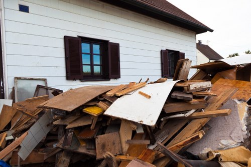 A large pile of mixed wooden debris and discarded construction materials is situated against the exterior wall of a white residential building with dark brown window shutters. The debris includes broken and weathered planks, sheets of particleboard, and various leftover wood pieces, some with nails or screws visible, scattered unevenly across the ground. The pile extends along the side of the building, partially covering a paved surface or driveway. In the background, additional rooftops and sky are visible, indicating a suburban or urban residential area. The lighting appears natural, likely daylight, highlighting the texture and aged appearance of the materials. The scene exemplifies typical outdoor waste accumulation from a property requiring garden clearance or rubbish removal services, as offered by Garden Clearance Brixton in the Brixton area postcode, with visual elements suggesting the need for efficient rubbish removal service to clear the debris from the property exterior.