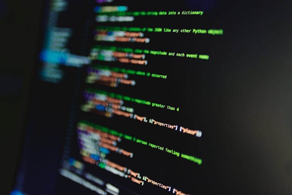 A close-up image of a person's hand typing on the keyboard of a silver laptop placed on a wooden surface. The person's wrist is adorned with a black watch, and the screen displays lines of code in a t