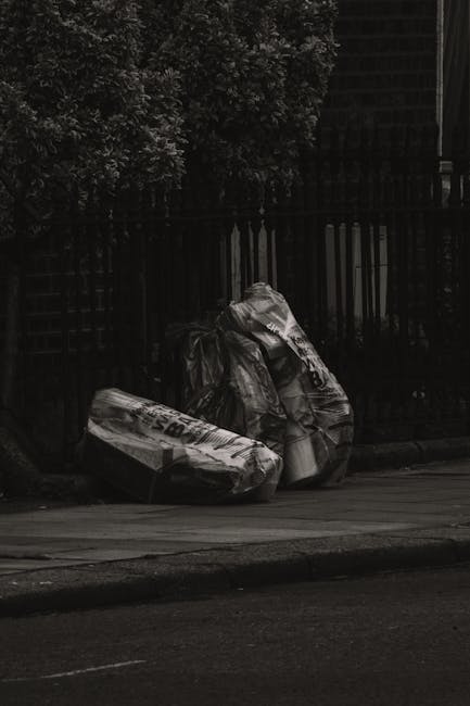 A large pile of waste and rubbish overflow from multiple bins located on a paved pavement area, with some waste spilling onto the ground. The collection includes various materials such as cardboard bo
