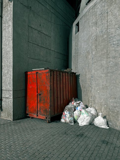 The image depicts a cluttered outdoor waste disposal area on a paved surface, featuring a large metal recycling bin overflowing with mixed paper, cardboard boxes, plastic wrapping, and other general w